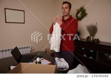 Man Folding Clothes At Home Office Desk With Christmas Decor And Laptop 134244168
