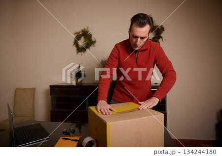 Man In Red Sweater Unpacks Package At Home Desk Amid Holiday Decor 134244180