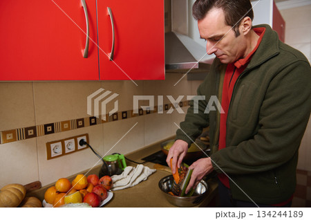 Man Grating Vegetables in Modern Home Kitchen With Red Cabinets and Fresh Produce Man Grating Vegetables in Modern Home Kitchen With Red Cabinets and Fresh Produce 134244189