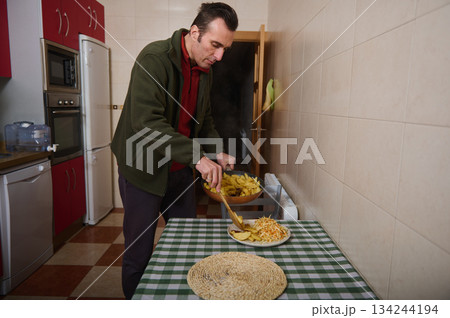 Man Serves Home-Cooked Dinner in Kitchen, Plating Potatoes and Fries on Checkered Table Man Serves Home-Cooked Dinner in Kitchen, Plating Potatoes and Fries on Checkered Table 134244194