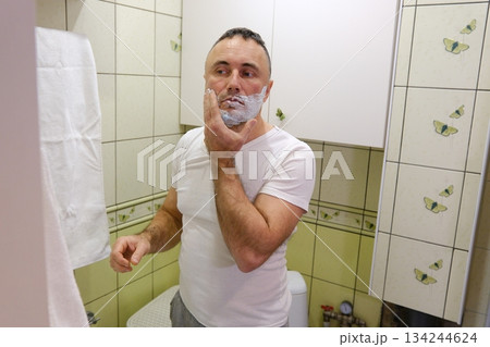a man shaves his beard at home in front of a mirror, men's cosmetics, shaving foam and lotion a man shaves his beard at home in front of a mirror, men's cosmetics, shaving foam and lotion 134244624