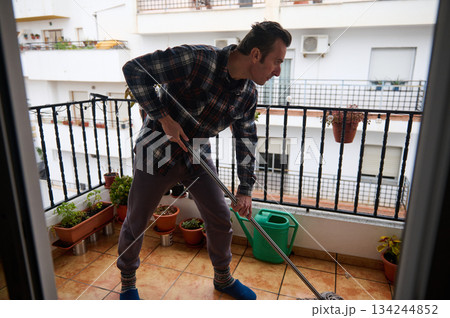 Man Mopping Balcony On Urban Apartment With Plants And Watering Can 134244852