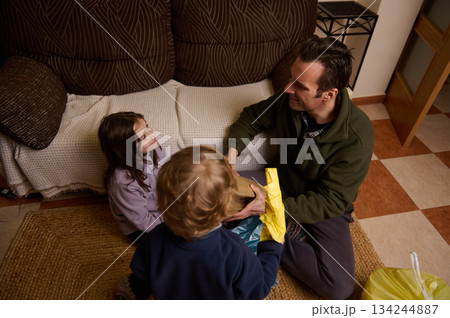 Father Shares Gift With Kids On Living Room Floor During Cozy Home Evening Father Shares Gift With Kids On Living Room Floor During Cozy Home Evening 134244887