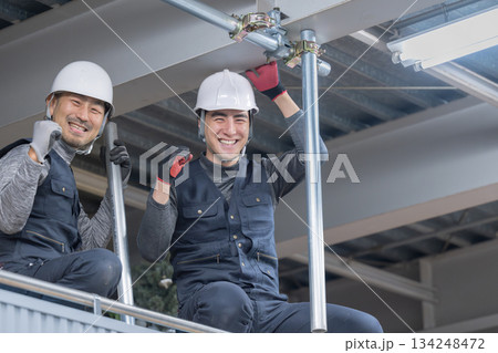 天井が近い高所の足場で単管パイプを固定するクランプなどの施工作業　鳶職や作業員のイメージ　笑顔でカメ 134248472