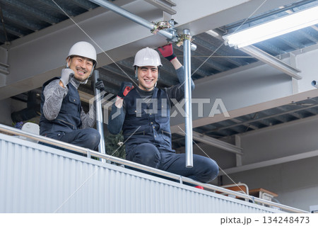 天井が近い高所の足場で単管パイプを固定するクランプなどの施工作業　鳶職や作業員のイメージ　笑顔でカメ 134248473