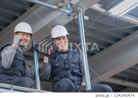 天井が近い高所の足場で単管パイプを固定するクランプなどの施工作業　鳶職や作業員のイメージ　笑顔でカメ 134248474