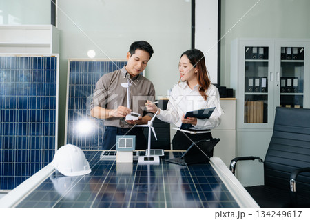 Two Asian engineers collaborate on a solar and wind energy project, discussing innovation and sustainability in a modern office setting. Two Asian engineers collaborate on a solar and wind energy project, discussing innovation and sustainability in a modern office setting. 134249617