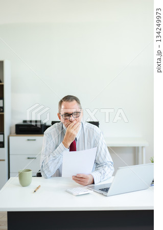 Focused senior executive analyzing printed documents at modern office desk with laptop and coffee, working on business strategy, finance, 134249973