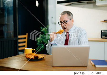 Mature Businessman using laptop while sitting at home. man sitting in kitchen and doing market research working Mature Businessman using laptop while sitting at home. man sitting in kitchen and doing market research working 134250055