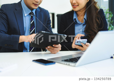Business documents on office table with smart phone and laptop and two colleagues discussing data in the background 134250789