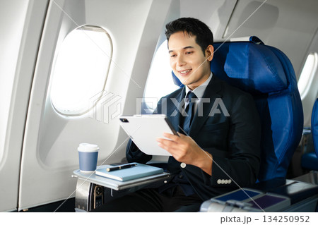 Confident businessman enjoying luxury air travel in first class, working on a tablet with a coffee, blending business, technology, and global connectivity. Confident businessman enjoying luxury air travel in first class, working on a tablet with a coffee, blending business, technology, and global connectivity. 134250952