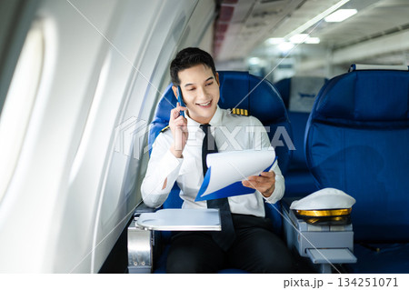 Confident airline pilot in uniform using wireless earbuds while reviewing documents, perfect for aviation, travel, and modern communication themes Confident airline pilot in uniform using wireless earbuds while reviewing documents, perfect for aviation, travel, and modern communication themes 134251071