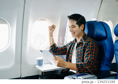 Happy young traveler celebrating victory with a fist pump on the airplane. Business success, achievement, and excited travel journey. Win, style, phone, tech. 134251189