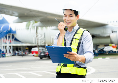 Ground crew engineer in safety vest and hard hat using walkie talkie while checking documents, perfect for aviation, maintenance, 134251237