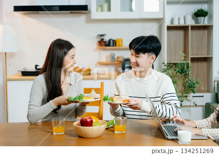 Asian parents toast sandwiches while daughter types on laptop during breakfast. A joyful mix of food, family bonding 134251269