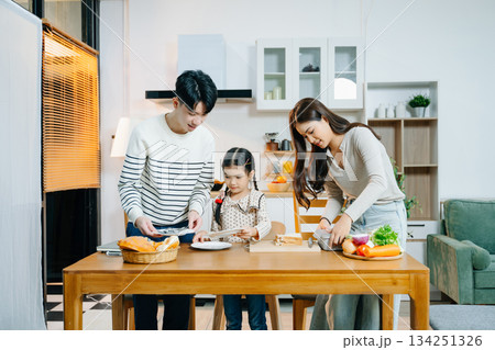 Asian family shares a happy morning, preparing and enjoying breakfast in a cozy kitchen. Parents and daughter bond over love Asian family shares a happy morning, preparing and enjoying breakfast in a cozy kitchen. Parents and daughter bond over love 134251326