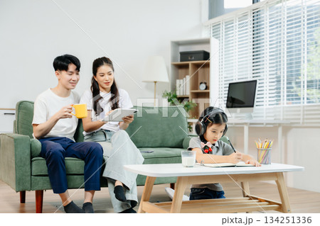 Asian child wearing headphones, studying and drawing at home, while parents enjoy coffee and tablet nearby. Family time, learning 134251336