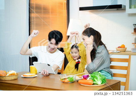 Happy Asian family cheers while using a tablet in the kitchen. Concept of digital learning, online success, family bonding, parenting, and joyful home moments. Happy Asian family cheers while using a tablet in the kitchen. Concept of digital learning, online success, family bonding, parenting, and joyful home moments. 134251363