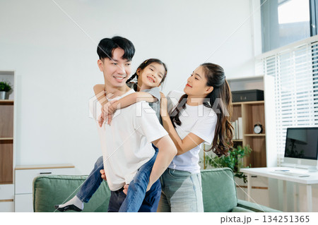 Asian parents and daughter enjoying a playful piggyback ride, laughing and bonding in cozy living room. Love, happiness, and family time Asian parents and daughter enjoying a playful piggyback ride, laughing and bonding in cozy living room. Love, happiness, and family time 134251365