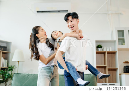Asian parents and daughter enjoying a playful piggyback ride, laughing and bonding in cozy living room. Love, happiness, and family time Asian parents and daughter enjoying a playful piggyback ride, laughing and bonding in cozy living room. Love, happiness, and family time 134251366
