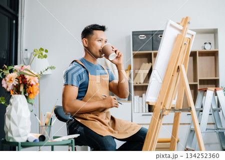Asian male painter do artwork in art workshop, painting supplies, oil pastels, two canvas easel, creative space with paintbrush in art studio 134252360