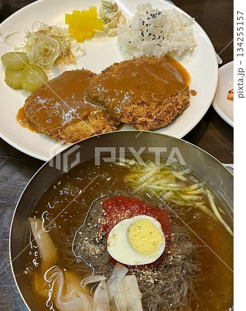 Cold naengmyeon noodles served in a chilled broth with sliced vegetables and egg, paired with crispy golden donkatsu, creating a balance of refreshing and savory flavors in one meal Cold naengmyeon noodles served in a chilled broth with sliced vegetables and egg, paired with crispy golden donkatsu, creating a balance of refreshing and savory flavors in one meal 134255157