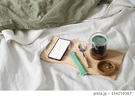 Smartphone, green superfood drink and vitamins on wooden tray in bed. Smartphone, green superfood drink and vitamins on wooden tray in bed. 134259367