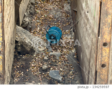 Deep excavation trench featuring robust concrete shoring walls and a discarded blue industrial hose reel, symbolizing ongoing urban utility infrastructure work captured by a mobile phone Deep excavation trench featuring robust concrete shoring walls and a discarded blue industrial hose reel, symbolizing ongoing urban utility infrastructure work captured by a mobile phone 134259597