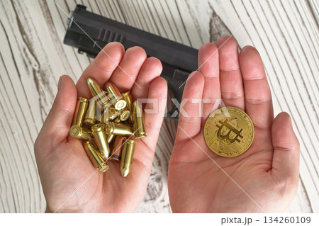 Yellow golden coloured bullets and bitcoin coins in two hands, blurred wooden desk with black gun under Yellow golden coloured bullets and bitcoin coins in two hands, blurred wooden desk with black gun under 134260109