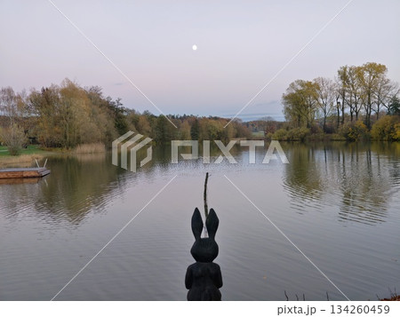 serene wooden rabbit sculpture overlooking calm lake with autumn trees reflected and moon overhead serene wooden rabbit sculpture overlooking calm lake with autumn trees reflected and moon overhead 134260459