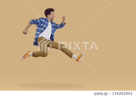 Happy brunette young man in casual clothes jumping in air on beige background, side view. 134262001