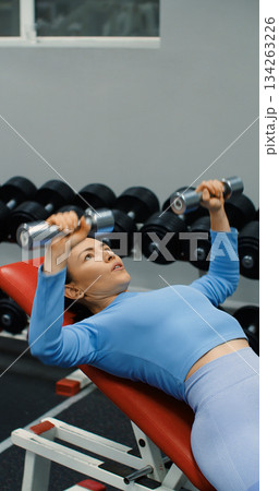 Close-up view of a fit Caucasian woman during gym workout. Bench press with dumbbells for chest. Gym scene shows woman performing bench press to tone chest and upper body. 134263226