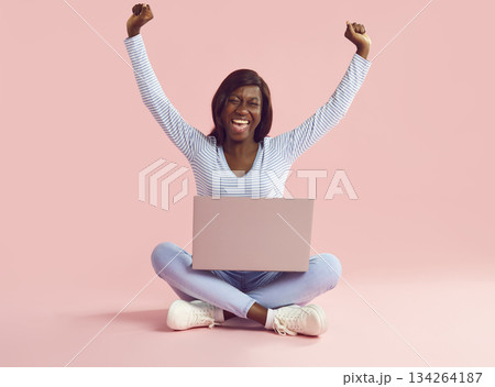 Woman sitting with a laptop with hands up in fists expressing happiness on studio pink background. Woman sitting with a laptop with hands up in fists expressing happiness on studio pink background. 134264187