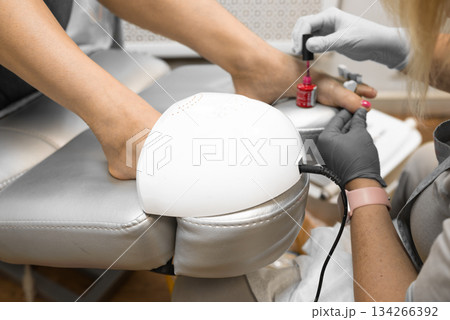 pedicure master applies gel polish to a woman toenails during procedure in a beauty salon. UV lamp for drying nail polish and gel 134266392