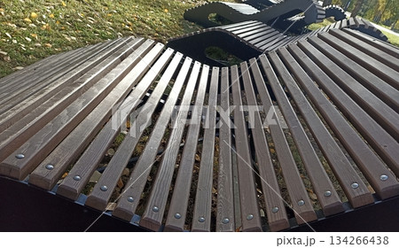 A modern wooden bench in a park provides a perfect spot for relaxation. This stylish outdoor seating enhances the landscape, inviting visitors to enjoy nature on a sunny day. 134266438