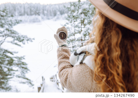 Woman in a hat holding a compass lying in hand on mitten against backdrop of snowy winter forest. Woman in a hat holding a compass lying in hand on mitten against backdrop of snowy winter forest. 134267490