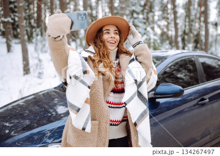 Young tourist takes a selfie with her phone next to her car on a snowy road. Travel concept. 134267497
