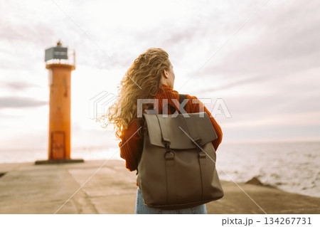 Woman enjoying breezy day near vibrant red lighthouse at sunset. Travel, weekend, lifestyle concept. 134267731