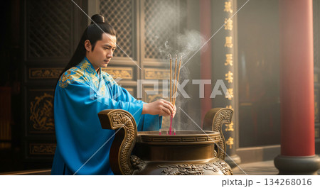 A man in traditional attire places joss sticks in a bronze container. The incense sends thin trails of smoke into the air inside a temple, creating a sacred atmosphere. A man in traditional attire places joss sticks in a bronze container. The incense sends thin trails of smoke into the air inside a temple, creating a sacred atmosphere. 134268016