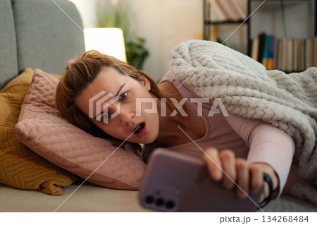 Woman experiencing shock while looking at phone in bed Woman experiencing shock while looking at phone in bed 134268484