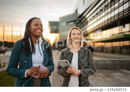 Diverse businesswomen smiling walking together outside modern office building Diverse businesswomen smiling walking together outside modern office building 134268552