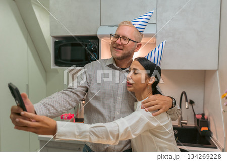 Couple Birthday Selfie Kitchen Celebration: Happy mature couple taking selfie photo in kitchen wearing party hats during birthday celebration. Couple Birthday Selfie Kitchen Celebration: Happy mature couple taking selfie photo in kitchen wearing party hats during birthday celebration. 134269228