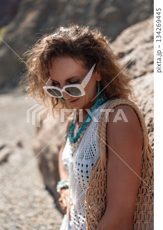 Woman, sunglasses, curly hair. Stylish woman with white shades and macrame bag standing outdoors, evoking a relaxed summer mood. Woman, sunglasses, curly hair. Stylish woman with white shades and macrame bag standing outdoors, evoking a relaxed summer mood. 134269445
