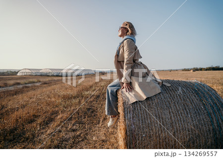 Woman in a Trench Coat Sitting on a Hay Bale in a Field Woman in a Trench Coat Sitting on a Hay Bale in a Field 134269557
