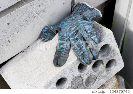 Single gray work glove with blue stripes lies draped over rough concrete block. White concrete walls visible in background. Light highlights texture of gloves and blocks, pause in hard work Single gray work glove with blue stripes lies draped over rough concrete block. White concrete walls visible in background. Light highlights texture of gloves and blocks, pause in hard work 134270746