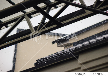 Heavy rain pouring off a black tiled roof and metal gutter below a modern, transparent canopy structure 134270920