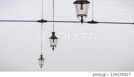 Three vintage street lamps hanging from wires against a bright, misty sky background Three vintage street lamps hanging from wires against a bright, misty sky background 134270927