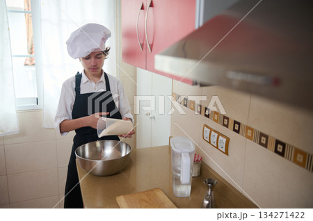 Young Chef In White Hat And Apron Measuring Flour In A Bright Home Kitchen 134271422