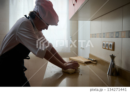 Chef Kneading Dough In Kitchen While Wearing White Coat And Hat 134271441