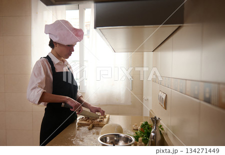 Young Chef In A Pink Toque Chops Mushrooms On A Cutting Board In A Bright Kitchen Young Chef In A Pink Toque Chops Mushrooms On A Cutting Board In A Bright Kitchen 134271449